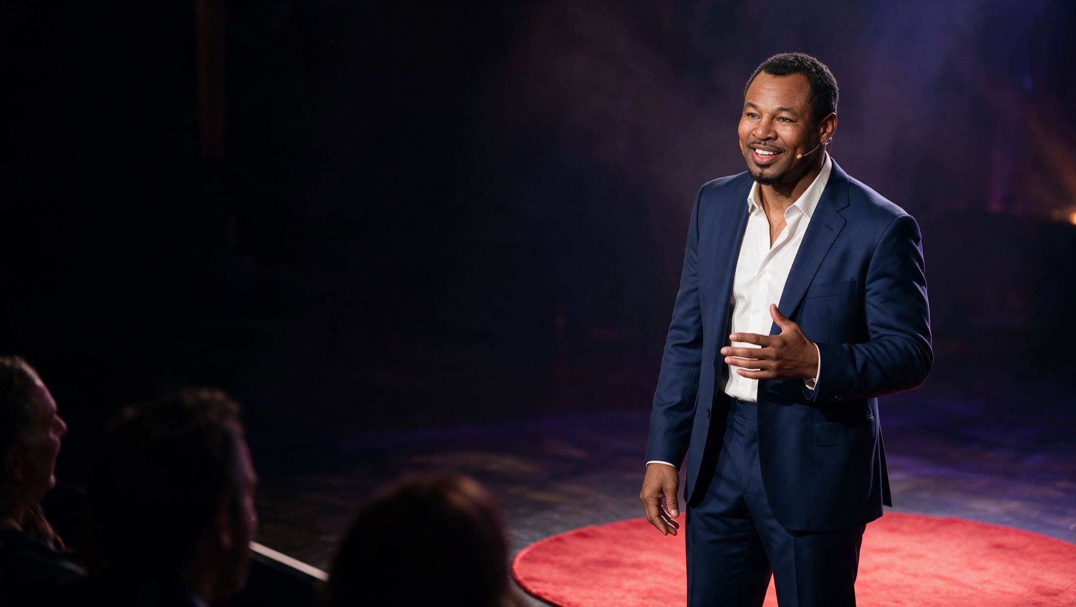 Sugar Shane Mosley giving a talk on stage, smiling, with a headset microphone, in front of an audience